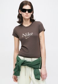 Nő fekete napszemüvegben, barna Nike pólóban, fehér nadrágban, zöld pulóverrel a derekán áll, fehér háttér előtt.