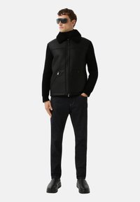 Zwart lederen gilet met een bontkraag, geribbelde zwarte mouwen, ritszakken op de borst, donkerblauwe spijkerbroek en zwarte stevige schoenen.