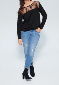 Schwarzes Langarmoberteil mit durchsichtigen Spitzenakzenten am Ausschnitt, kombiniert mit hellblauen, verwaschenen Jeans und schwarzen Rauleder-Ankle-Boots mit Blockabsatz.
