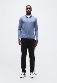 Άνδρας που στέκεται κρατώντας ένα μπαστούνι του γκολφ, φοράει μπλε φαρδιά μπλούζα με φερμουάρ, μαύρο παντελόνι και λευκά παπούτσια γκολφ σε απλό φόντο.