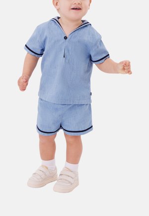 Kleinkind trägt hellblaues kurzärmeliges Hemd und passende Shorts mit dunklem Rand, weiße Socken und beige Klettverschluss-Sneaker, steht vor weißem Hintergrund.