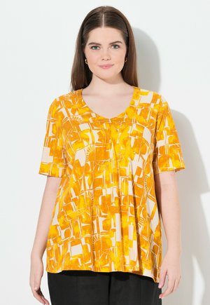 Haut jaune à motifs avec un col en V et des manches courtes, présentant des formes et textures abstraites dans un tissu léger. Associé à un pantalon sombre.