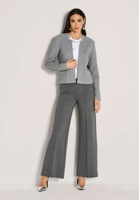 Vrouw in een grijze herringbone blazer, witte top, grijze wijde broek, zilveren hoge hakken en druppelvormige pareloorbellen, staande tegen een lichte muur.