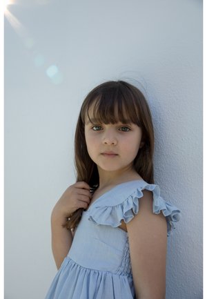 Mladé dievča s dlhými hnedými vlasmi v svetlomodrých šatách s volánikmi, stojace pred bielou textúrovanou stenou, pokojne sa pozerajúce do kamery.