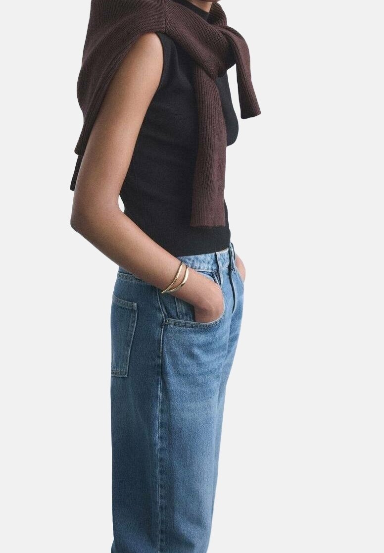 Personne portant un haut noir sans manches avec un pull marron posé sur les épaules et les mains dans les poches d'un jean bleu, bracelets en or au poignet.