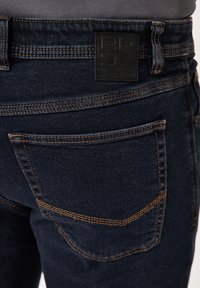 Dark blue denim jeans feature a textured black leather patch with logo on the waistband, contrasting orange stitching, and back pocket design.