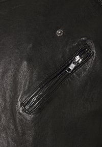 Svart lädertextil med en slät yta, med en dragkedjeficka och en tryckknappdetalj. Dragkedjan är i metall med ett rektangulärt handtag.