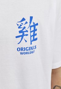 Camiseta de algodón blanca con un gráfico en negrita en azul que incluye símbolos y las palabras "ORIGINALS WORLDWIDE". Diseño de mangas cortas y cuello redondo.