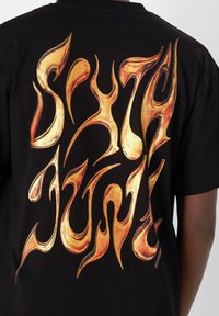Camiseta negra con texto dorado llameante en un diseño abstracto y similares a llamas en la parte posterior. Mangas cortas y cuello redondo clásico.