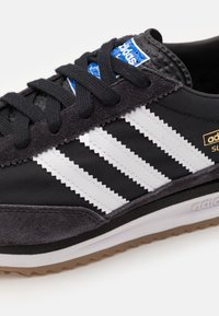 Svart och grå atletisk sko med tre vita tandade ränder, textil- och mockamaterial, svarta snörningar och en gummigummisula.