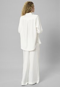 Frau mit lockigem Haar, die ein lockeres weißes Langarmshirt und weite weiße Hosen trägt, steht vor einem schlichten grauen Hintergrund.