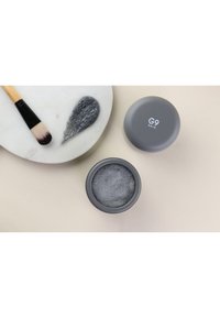 Un pot circulaire gris étiqueté "G9 SKIN" avec une crème noire texturée visible à l'intérieur, accompagné d'un pinceau noir et d'une traînée grise et mousseuse.