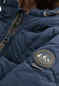 Marineblauer Steppjacke mit strukturierten Oberflächen. Verfügt über einen Reißverschluss, ein braunes Innenfutter und ein rundes Logo-Patch am Ärmel.