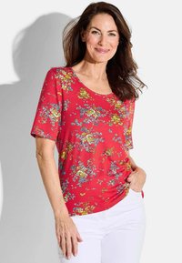 Rotes Blusenshirt mit kurzen Ärmeln und rundem Ausschnitt, das eine Mischung aus gelben und blauen Blumenmustern auf einem glatten Stoff zeigt.