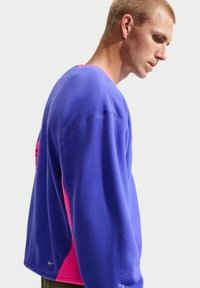 Νεαρός άνδρας με μπλε και ροζ φλις πουκάμισο με μακριά μανίκια κοιτάζει προς τα κάτω, μπροστά σε απλό ανοιχτό φόντο.