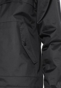 Zwarte waterdichte jas met een zijzak met knopen, een slanke afwerking en een getextureerde stof voor duurzaamheid en weerbestendigheid.