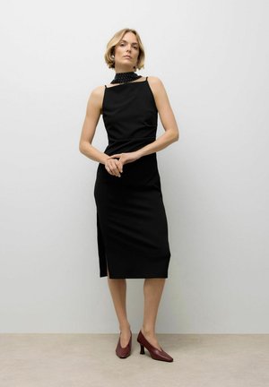 Crna midi haljina s tankim naramenicama i visokim ovratom s uzorkom točkica. Bočni prorez. Nosena s bordo cipelama na debljoj peti. Elegantno pristajanje.