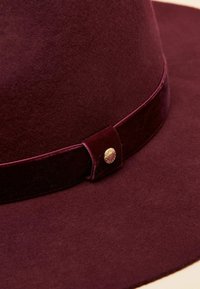 Pălărie din fetru burgundy, cu o bandă de catifea și un nasture auriu ca accent, textură netedă, bor larg și coroană structurată.