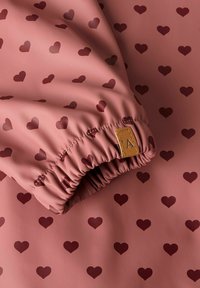 Tissu caoutchouté en rose poussiéreux avec un motif de cœurs rouge foncé. Comprend un poignet élastique et une étiquette beige avec la lettre "A."