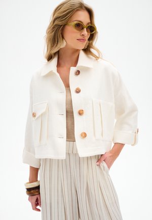 Kobieta ubrana w oversize białą zapinaną na guziki kurtkę, szerokie spodnie w paski, beżowy top, zielone okulary przeciwsłoneczne oraz masywne bransoletki, pozująca z jedną ręką w kieszeni.