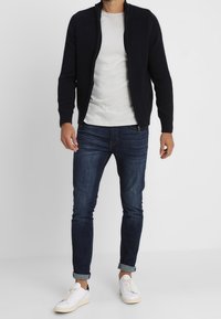 Námořnický zipový svetr s texturovaným pletením, bílý tričko s krátkým rukávem, tmavé džíny a bílé tenisky. Úzký střih s ohrnutými manžetami.