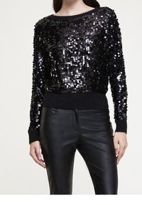 Svart paljettröja med rund halsringning, ribbade muddar och fåll. Matchad med svarta läderbyxor som framhäver en glänsande textur och en figurnära design.