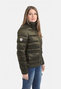 Veste matelassée vert olive avec un col haut, un design matelassé horizontal et des poches avant. Comprend un patch logo sur la manche gauche.