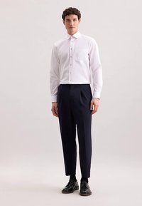 Camicia bianca a maniche lunghe con taschino sul petto, chiusura con bottoni, abbinata a pantaloni eleganti blu navy e scarpe nere. Tessuto liscio, vestibilità sartoriale.