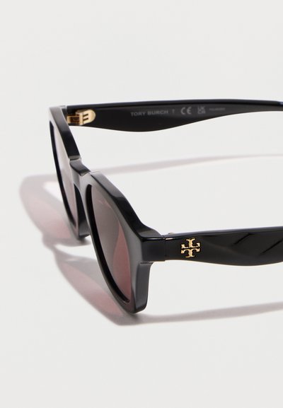 Μαύρα ορθογώνια γυαλιά ηλίου με σκοτεινούς φακούς και χρυσό λογότυπο Tory Burch στα πλαϊνά μπράτσα, παρουσιασμένα σε λευκό φόντο.
