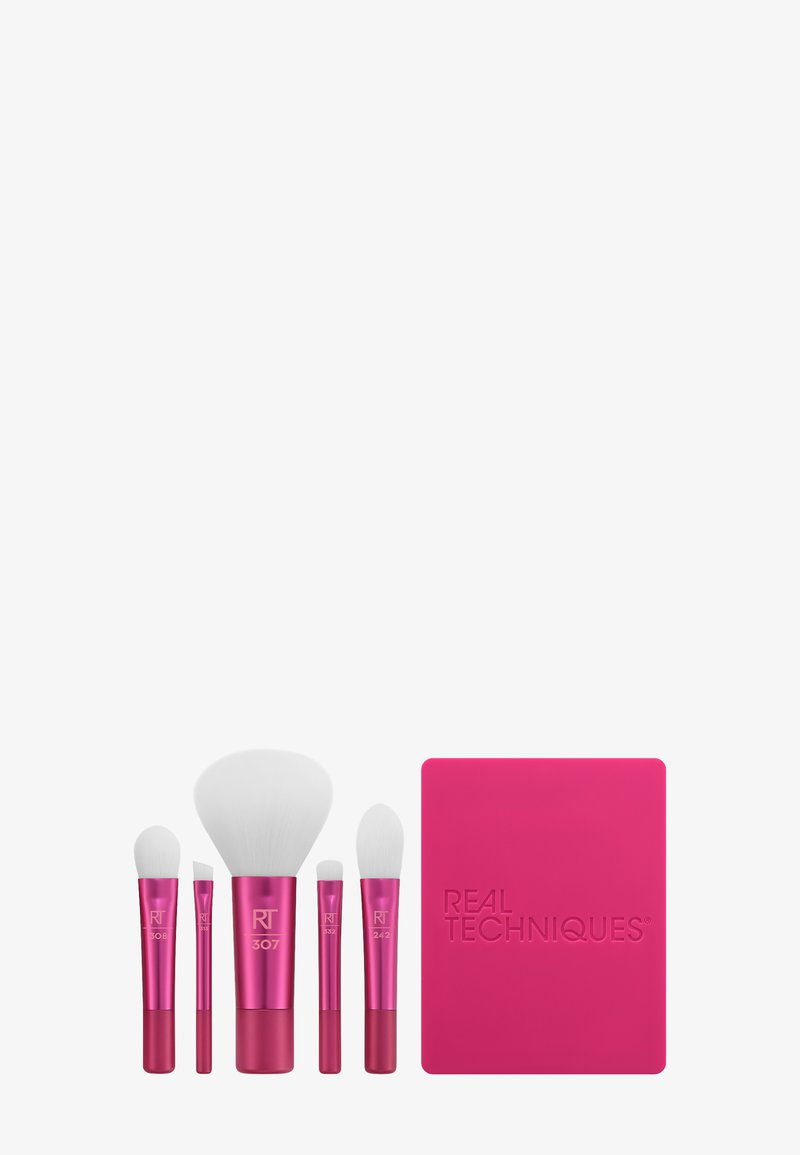 Cinq pinceaux de maquillage à manches roses avec des poils blancs, accompagnés d'un porte-pinceaux rectangulaire Real Techniques assorti de couleur rose.