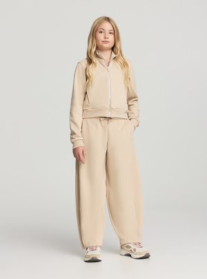 Giacca beige con zip e design corto, abbinata a pantaloni larghi coordinati. Tessuto morbido, abbinata a sneakers chiare.