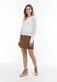 Bílý háčkovaný svetr s výstřihem do V, v kombinaci s hnědou mini sukní. Modelka má na sobě béžové šněrovací boty s robustní podrážkou. Hladké pozadí.
