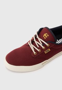 Sapatilha de lona cor bordô com atacadores crème, sola branca e pequena etiqueta "etnies" próxima aos ilhós, em fundo branco liso.