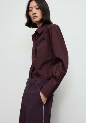 Camisa burdeos de botón, hecha de tejido texturizado, con mangas largas y cuello clásico, combinada con pantalones oscuros con ribete blanco.