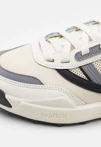 Sneaker caratterizzata da una miscela di pelle color crema, grigio e nera. Design strutturato con dettagli perforati, lacci piatti e un logo "adiPRENE" sulla suola.
