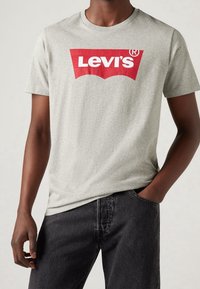 Harmaa puuvillainen T-paita, jossa suuri punainen Levi's-logo erottuvana kaarevana kuviona. Pyöreä kaula-aukkko ja lyhyet hihat, yhdistetty tummiin farkkuihin.