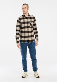 Flanellen shirt met ruitpatroon in beige, zwart en grijs, met knoopsluiting, een enkele borstzak, gecombineerd met blauwe jeans en sneakers.