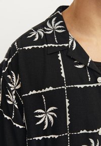 Camisa negra con un patrón de palmeras en blanco. Tiene un cuello clásico y botones, con una textura suave y mangas cortas.