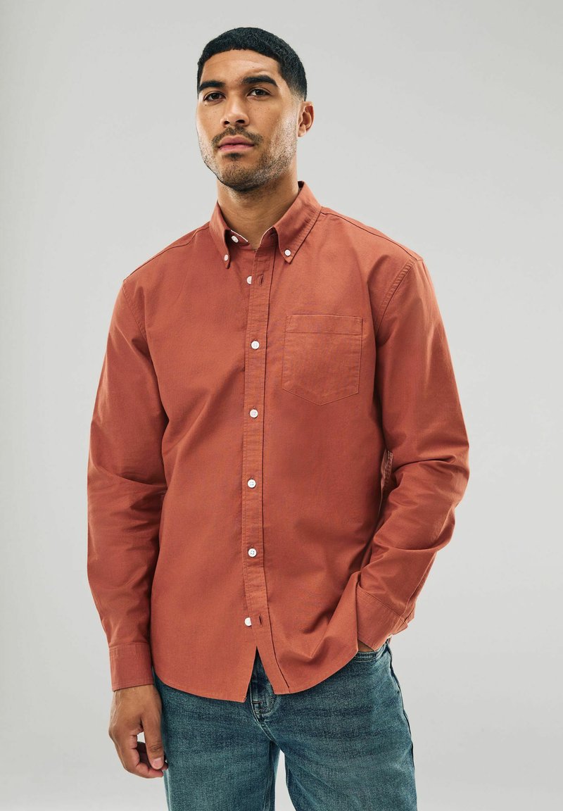 Rust-colored button-up shirt in cotton with a small chest pocket and white buttons, paired with blue jeans. Smooth texture, long sleeves.