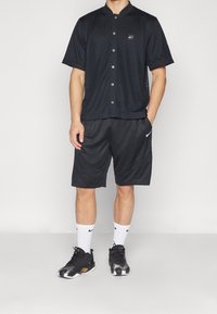 Camicia a maniche corte nera in rete con bottoni, abbinata a pantaloni corti neri e sneakers nere. Logo Nike bianco sulla camicia e sui calzini.