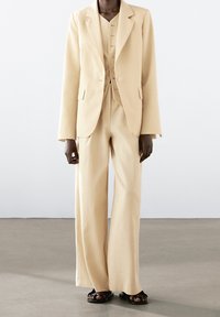 Beżowy, dopasowany garnitur z usztywnioną marynarką i szerokimi spodniami, z zapinanym na guziki kamizelką pod spodem. Gładka tkanina i minimalistyczny design.