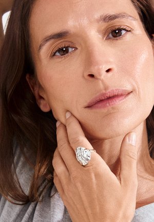 Žena s hnedými očami a vlasmi, ktorá si dotýka brady, nosí na prste veľký strieborný prsteň s abstraktným textúrovaným dizajnom.