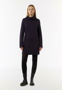 Lila gestricktes Sweaterkleid mit hohem Kragen, gerippten Bündchen und gerader Silhouette. Getragen mit schwarzen Strumpfhosen und klobigen schwarzen Stiefeln.