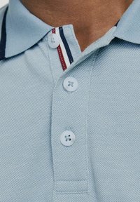 Ljusblå polotröja med textilstruktur, en krage med två knappar och marinblå, röd och vit randig detalj på kragen.