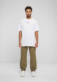 T-shirt blanc à manches courtes avec logo en script noir, associé à un pantalon ample vert olive et des baskets blanches. Tenue simple et décontractée.