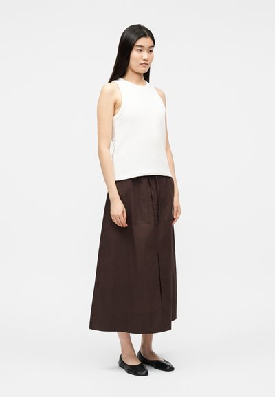 Femme aux longs cheveux noirs portant un haut blanc sans manches, une jupe midi brun foncé avec une fente à l'avant, et des chaussures plates noires, se tenant devant un fond blanc.