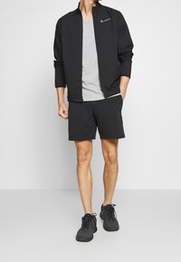 Černá sportovní bunda Nike se zipem, šedé tričko a černé kraťasy v kombinaci s černými teniskami, které představují sportovní design.