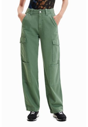 Personne portant un pantalon cargo vert olive avec de grandes poches latérales, des baskets noires et un haut sombre à motifs, les mains dans les poches.