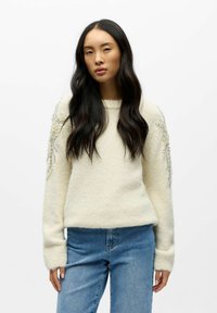 Weißer Strickpullover mit langen Ärmeln, der strukturierte Akzente und Verzierungen an den Schultern aufweist, kombiniert mit hellblauen Jeans.