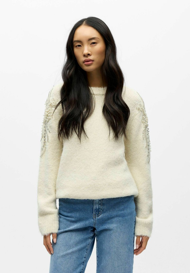 Weißer Strickpullover mit langen Ärmeln, der strukturierte Akzente und Verzierungen an den Schultern aufweist, kombiniert mit hellblauen Jeans.